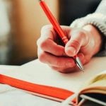 A student writing in a notebook with an orange pen.