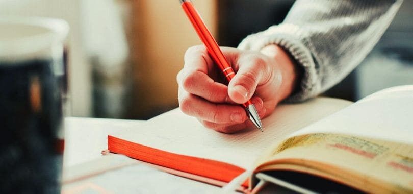 A student writing in a notebook with an orange pen.