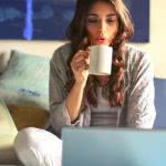 A college student sitting on her bed, drinking coffee, and studying on a laptop.