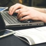 A student typing on a laptop with a booklet next to them.