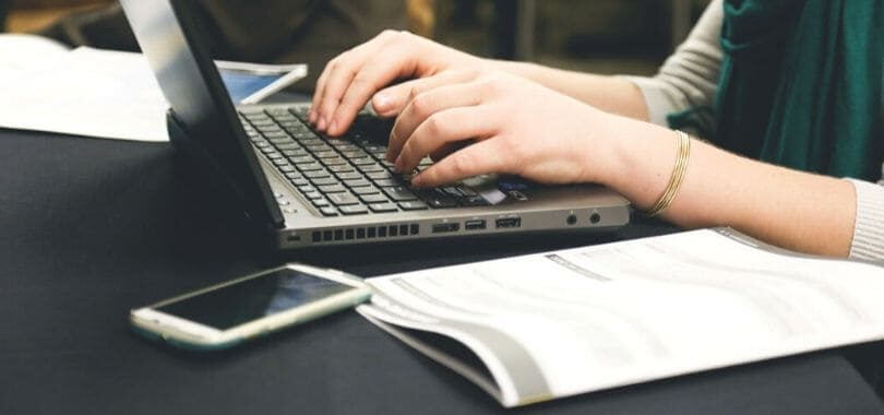 A student typing on a laptop with a booklet next to them.