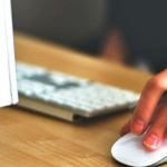 A student holding a mouse and a pen while sitting in front of a computer monitor.