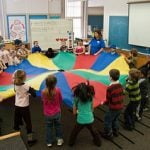 Kids gather around the big color wheel fabric and holding it.