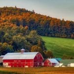 A red barn on grassy farmland with hills covered in trees behind it.