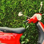 A red moped parked in front of a green hedge.