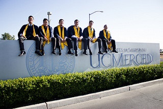 Students in graduation gowns sitting atop of the entrance sign at UC Merced.