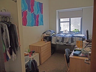 Dorm room with stuff inside such as clothes, desk, bag, and bed.