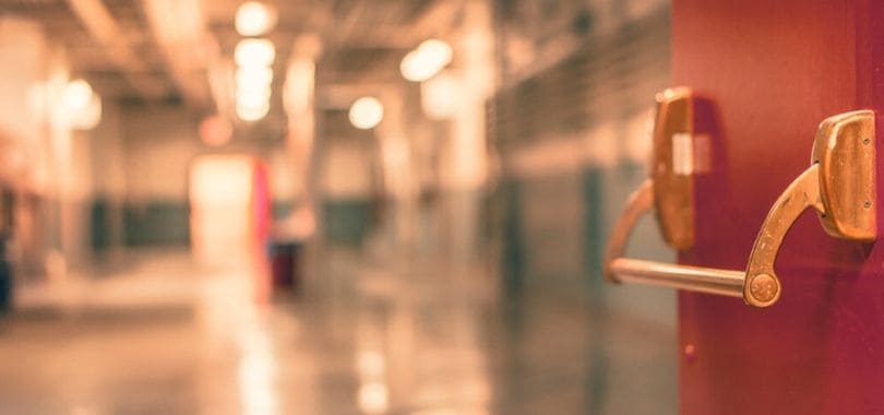 A red push door in a high school, opening up to a hallway with lockers.