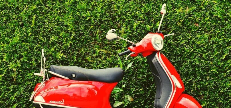 A red moped parked in front of a green hedge.