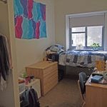 Dorm room with stuff inside such as clothes, desk, bag, and bed.