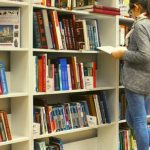 A college student standing in front of multiple bookshelves reading a book.