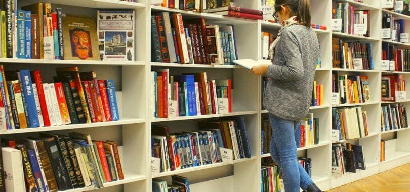 A college student standing in front of multiple bookshelves reading a book.
