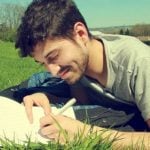 A student lying on a lawn writing down notes on a notebook.