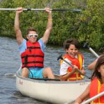 Camp counsellor holding oar above his head guiding children in kayaks on a lake.