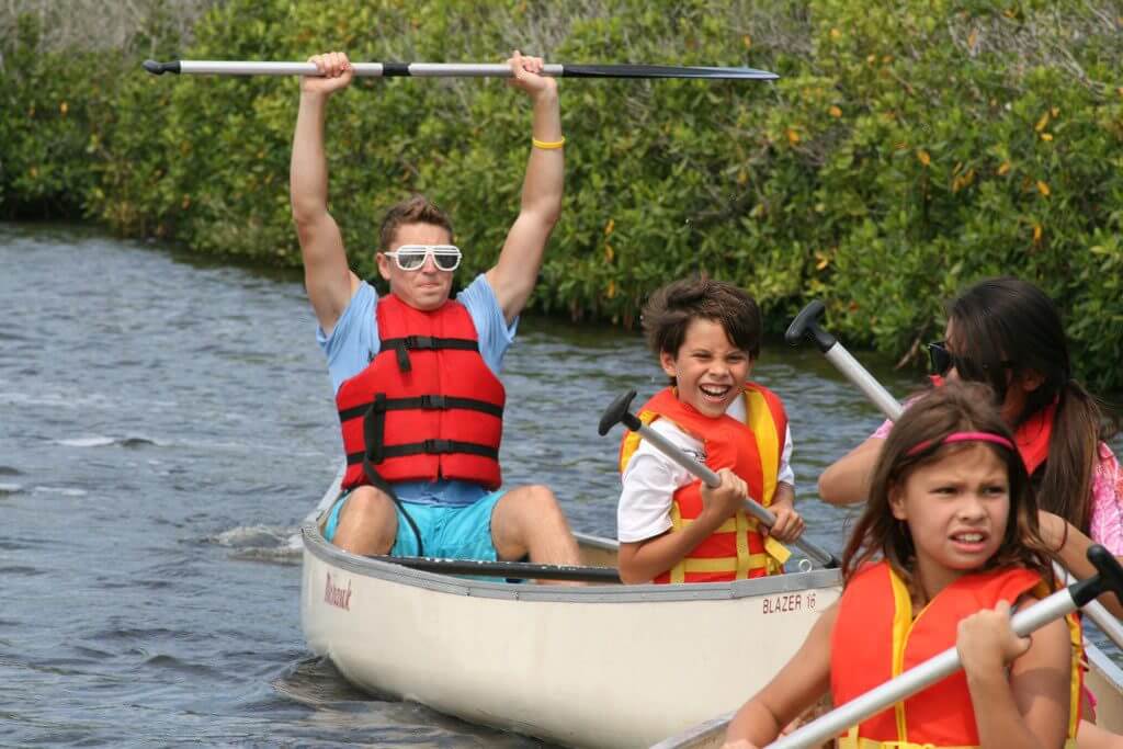 Camp counsellor holding oar above his head guiding children in kayaks on a lake.
