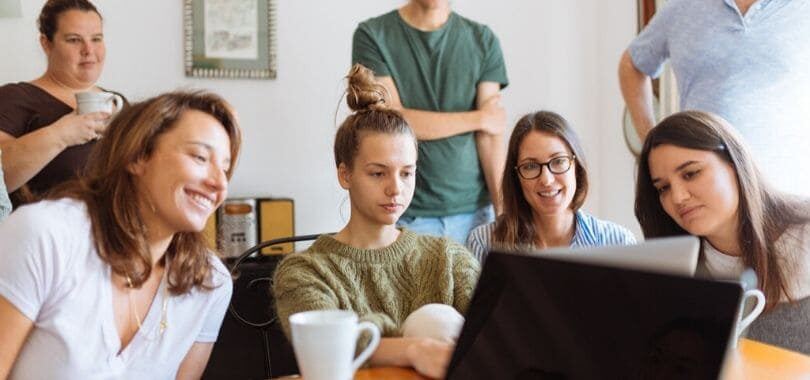 A group of students sitting together in front of one laptop, with a few students standing behind them.