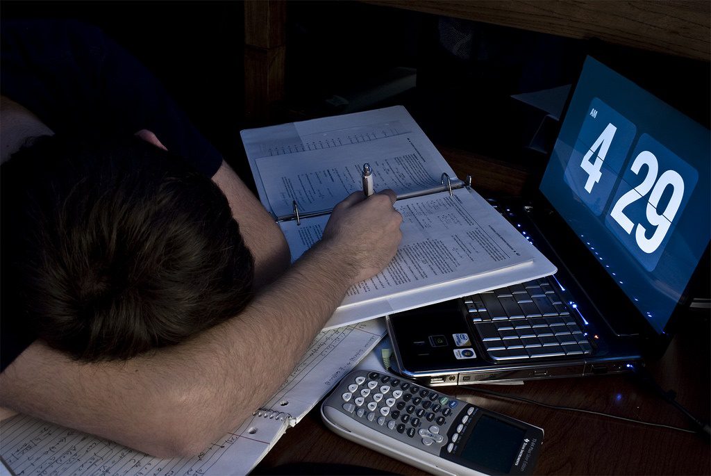 A male student fell asleep on the desk with papers and a laptop while holding a pen.
