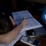 A male student fell asleep on the desk with papers and a laptop while holding a pen.