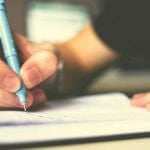 A person holding a blue pencil writing in a notebook.