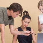 One student sitting at a laptop with two other people standing around them, with one pointing towards the laptop.