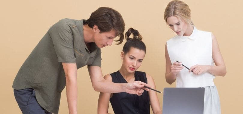 One student sitting at a laptop with two other people standing around them, with one pointing towards the laptop.
