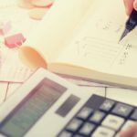 A student writing down expenses on a pad of paper with a calculator next to them.