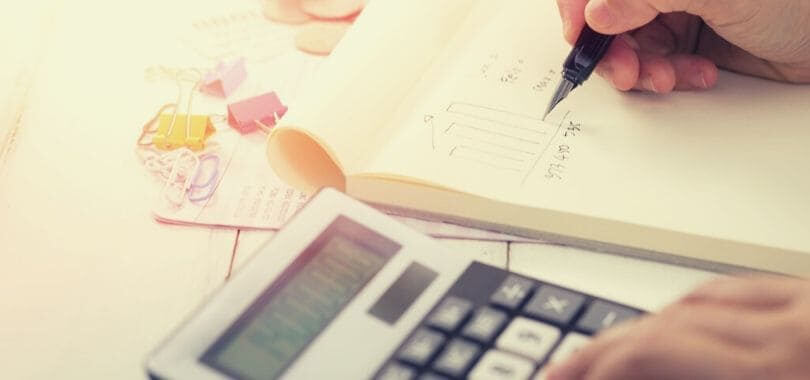 A student writing down expenses on a pad of paper with a calculator next to them.