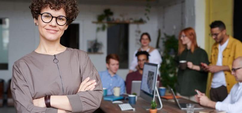 A person standing with crossed arms and smiling while more people are meeting in the background.