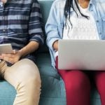 Two college students sitting together, one is holding a laptop and one is holding an iPad, shown from shoulders to knees.