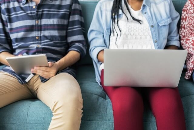 Two college students sitting together, one is holding a laptop and one is holding an iPad, shown from shoulders to knees.