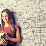 A student holding a stack of books, standing against a white board covered in writing.