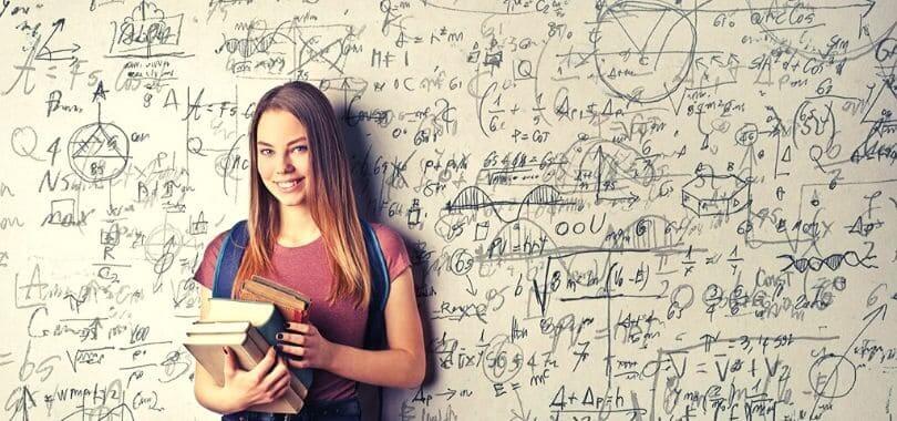 A student holding a stack of books, standing against a white board covered in writing.