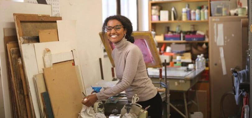 A student smiling, standing by a table.