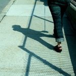 A student walking down the sidewalk with a visible shadow, shown from the waist down.