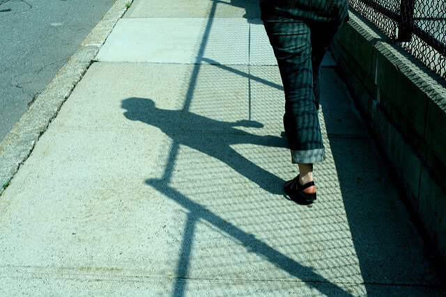 A student walking down the sidewalk with a visible shadow, shown from the waist down.