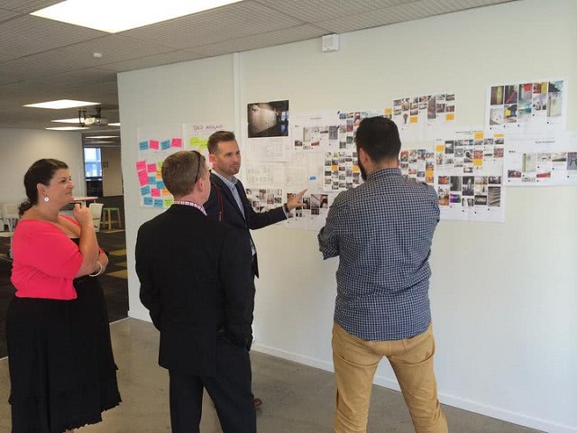 A man is pointing his hand on the whiteboard with images on it while discussing to three people.