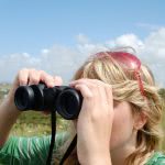 A blonde girl looking through binoculars in the field.