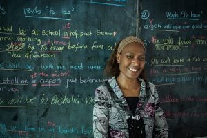 A female teacher is smiling in front of the green board with colorful chalks.