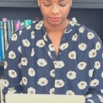 A college student sitting in front of a bookcase with a laptop in their lap.