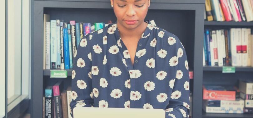 A college student sitting in front of a bookcase with a laptop in their lap.