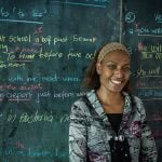 A female teacher is smiling in front of the green board with colorful chalks.