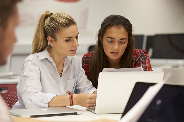 Two ladies are sitting on a desk while looking on the laptop.