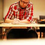 A male student wearing red checkered polo, writing on the desk.