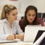 Two ladies are sitting on a desk while looking on the laptop.