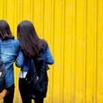 Two girls standing side by side against a yellow wall.