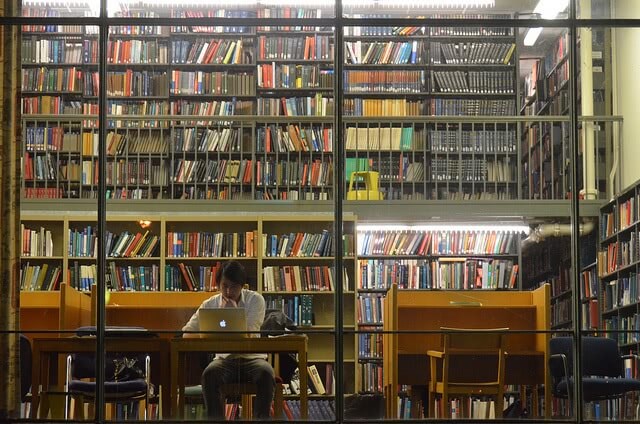 Man studying alone on his Macbook at the library showing bunch of books behind him.