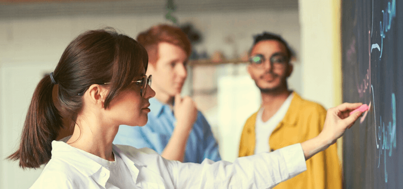 A tutor writing on a chalkboard with two students standing behind them.