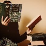 A girl covering her face with an open book while holding another book on the left hand.