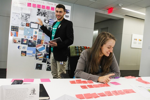 A woman sitting on a desk while looking at her notes and a man standing behind her discussing something.