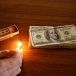 A hand lighting up a match beside a pile of a dollar bill.